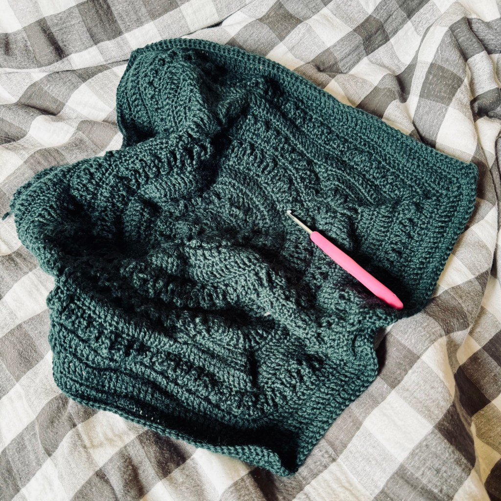 A pink-handled crochet hook rests on an intricate green crochet granny square, which is slightly rumpled on top of a slightly rumpled grey and white checked quilt cover.